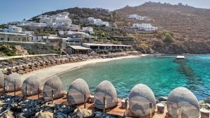 foto von strandkörben auf einer holzterrasse mit glaswand dahinter über einem felsenstrand mit blick auf den sandstrand mit sonnenliegen und auf das resort am hang mit weissen häusern und natürlich auf das kristallklare smaragd grüne türkise wasser des oz
